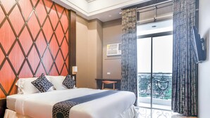 Ein Hotelzimmer mit einem großen Bett, einem Holzkopfteil, einem Balkon mit gemusterten Vorhängen und Blick auf die Stadt.