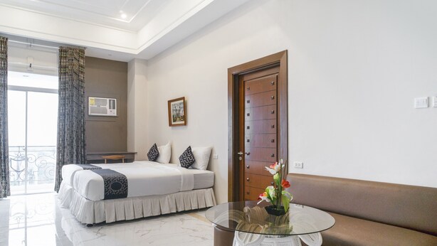 Ein Hotelzimmer mit einem Bett, einem runden Glastisch mit Pflanze, einer Bank, einer Tür und einem Balkon mit Vorhängen.