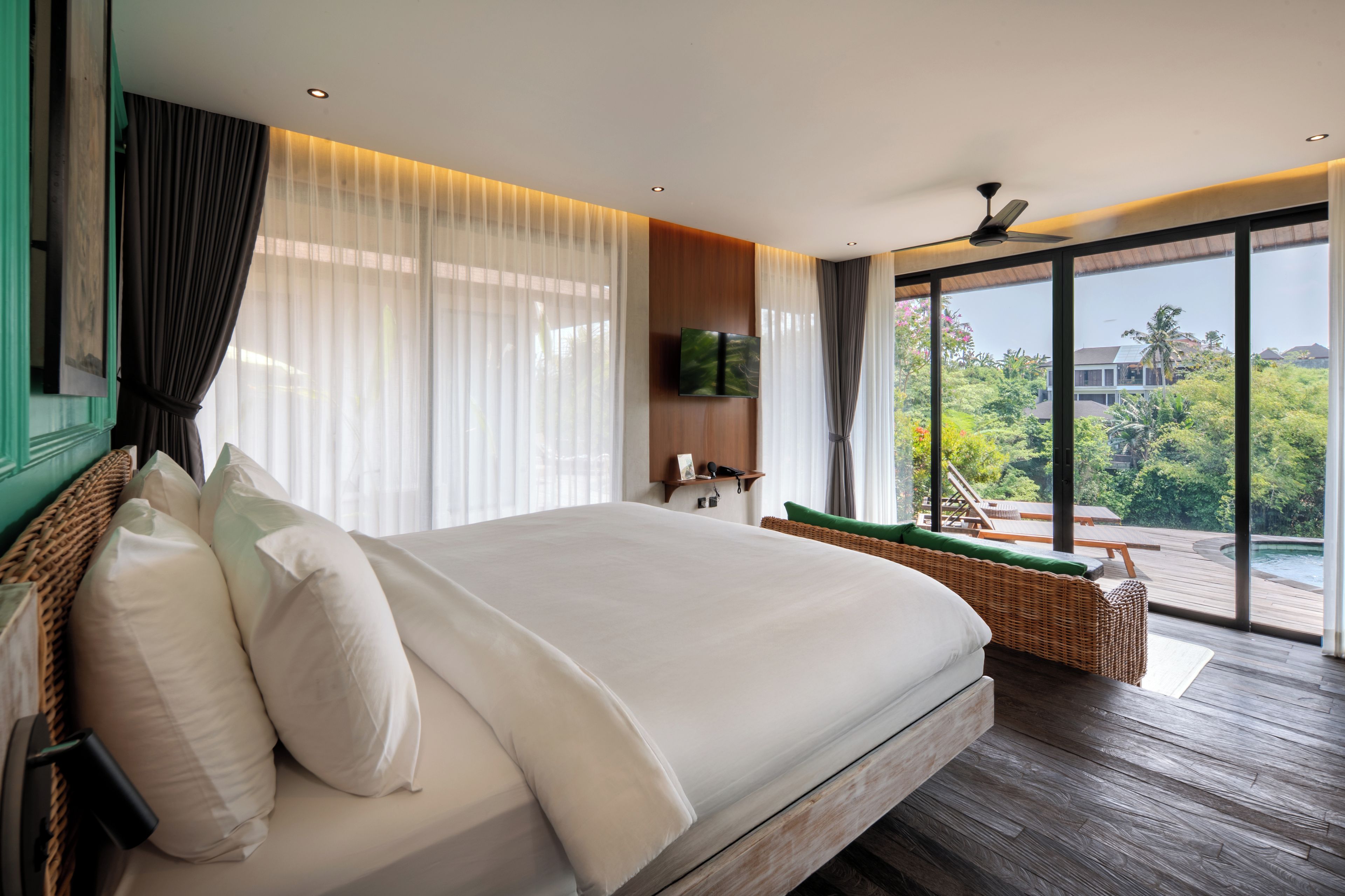 A spacious bedroom with a large bed, a television, and a view of a swimming pool and greenery.
