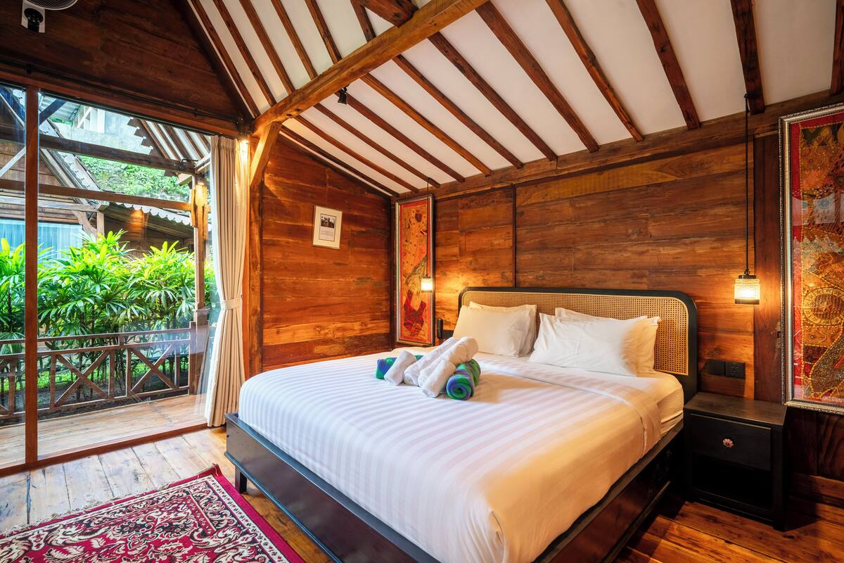 A wooden room with a bed, a nightstand, and a view of greenery outside.