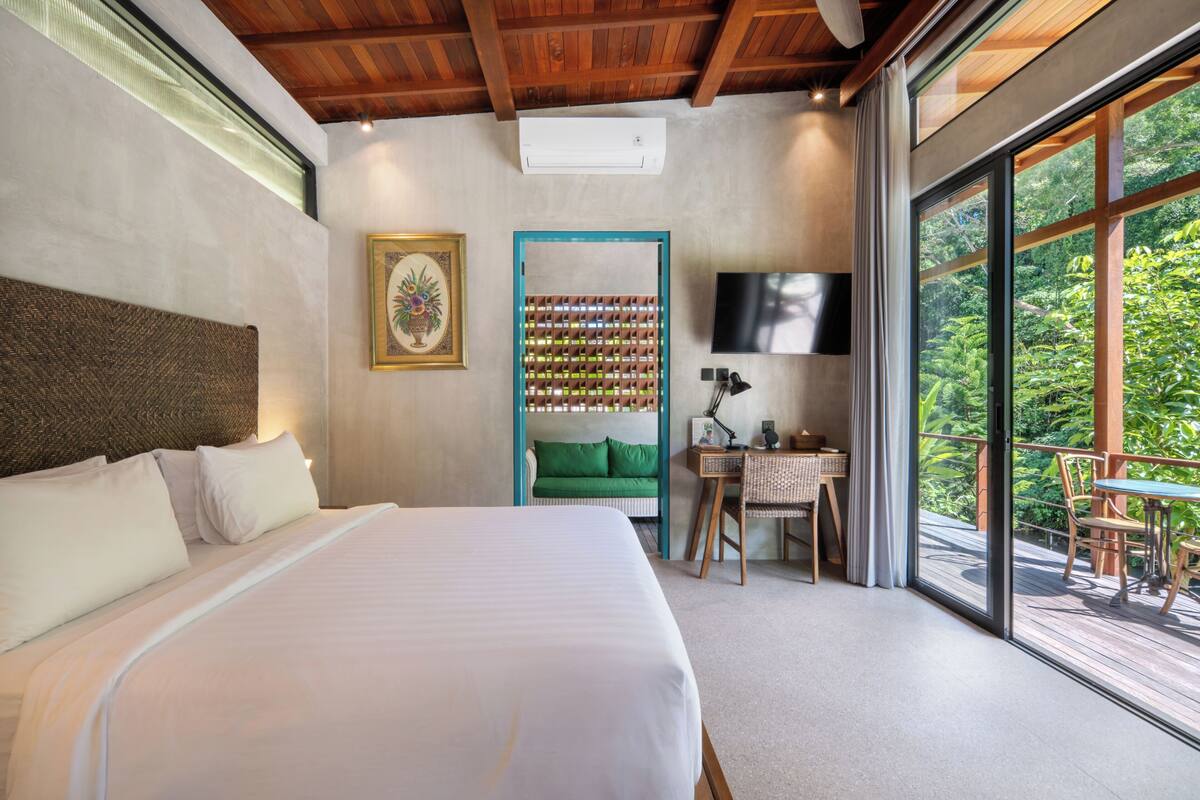 A bedroom with a large bed, a desk with a chair, a television, and a balcony with a view of greenery.