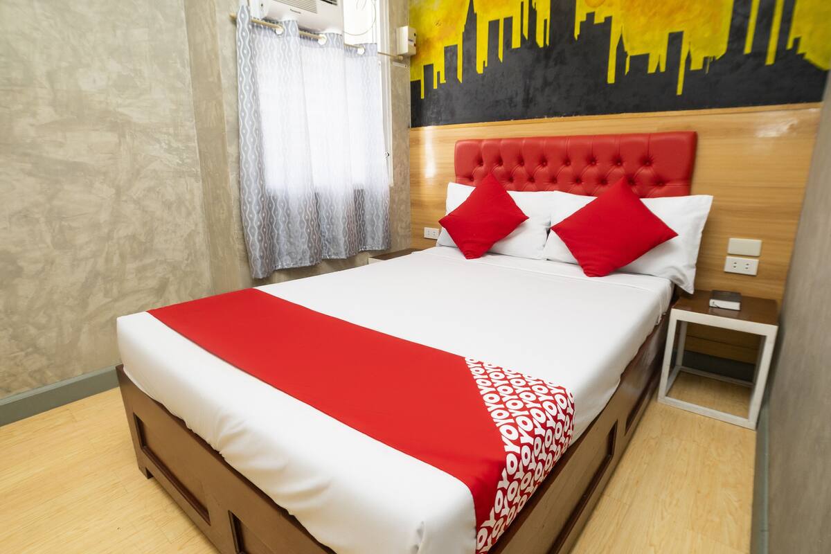 A hotel room with a large bed, a red and white bedspread, a city skyline mural on the wall, and a wooden headboard.