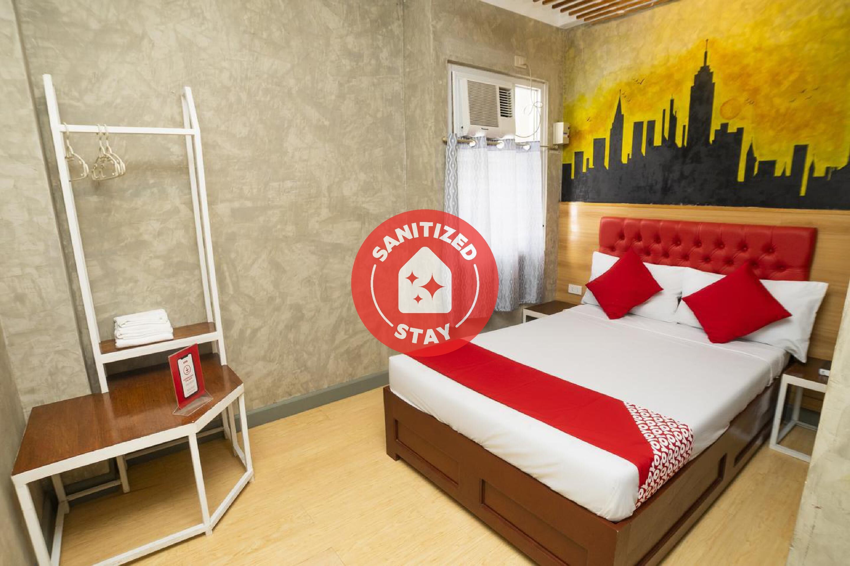 A hotel room with a bed, a white shelf with towels, a red and white bedspread, and a city skyline mural on the wall.