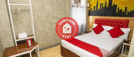 A hotel room with a bed, a white shelf with towels, a red and white bedspread, and a city skyline mural on the wall.