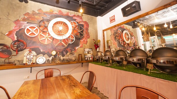 Un ristorante con un lungo tavolo di legno e sedie in metallo, un murale raffigurante ingranaggi e un orologio, e un'area buffet con piatti di servizio in metallo.