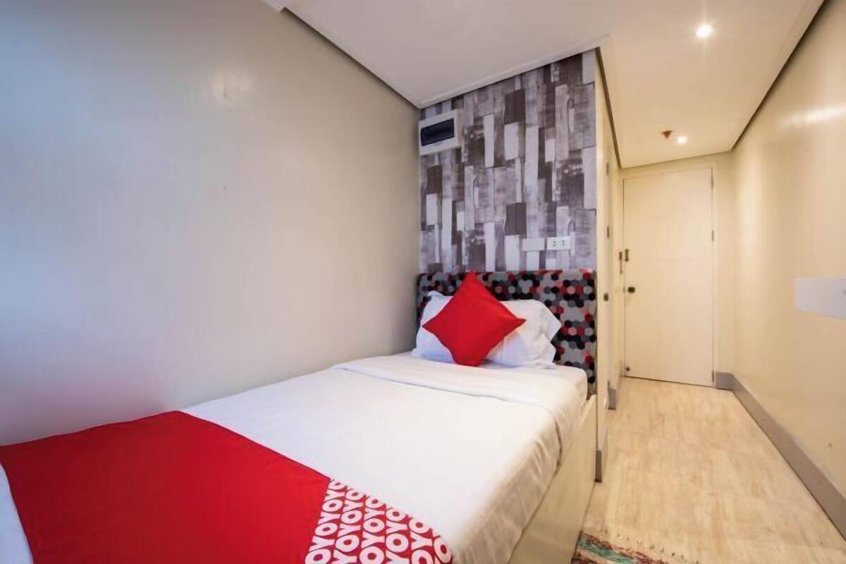 Ein kleines, ordentlich eingerichtetes Schlafzimmer mit einem Bett, einer roten und weißen Decke mit Muster, einer Wand mit geometrischem Design und einer Tür, die in einen anderen Raum führt.