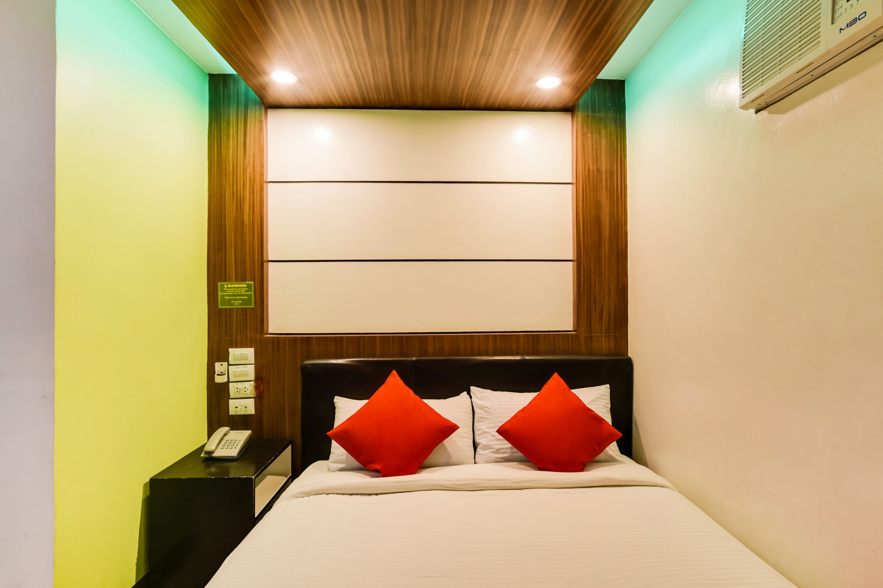 A hotel room with a wooden headboard, two red pillows, a white bedspread, and a nightstand with a telephone.