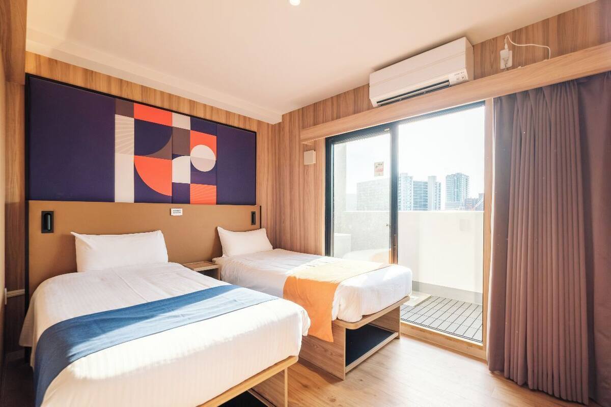 Ein Hotelzimmer mit zwei Betten, einem großen Fenster mit Blick auf die Stadt und einer Klimaanlage.