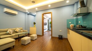 Un salon moderne avec un canapé, des poufs et une table basse en verre. On aperçoit un coin cuisine avec une cuisinière et des placards à travers une porte ouverte.