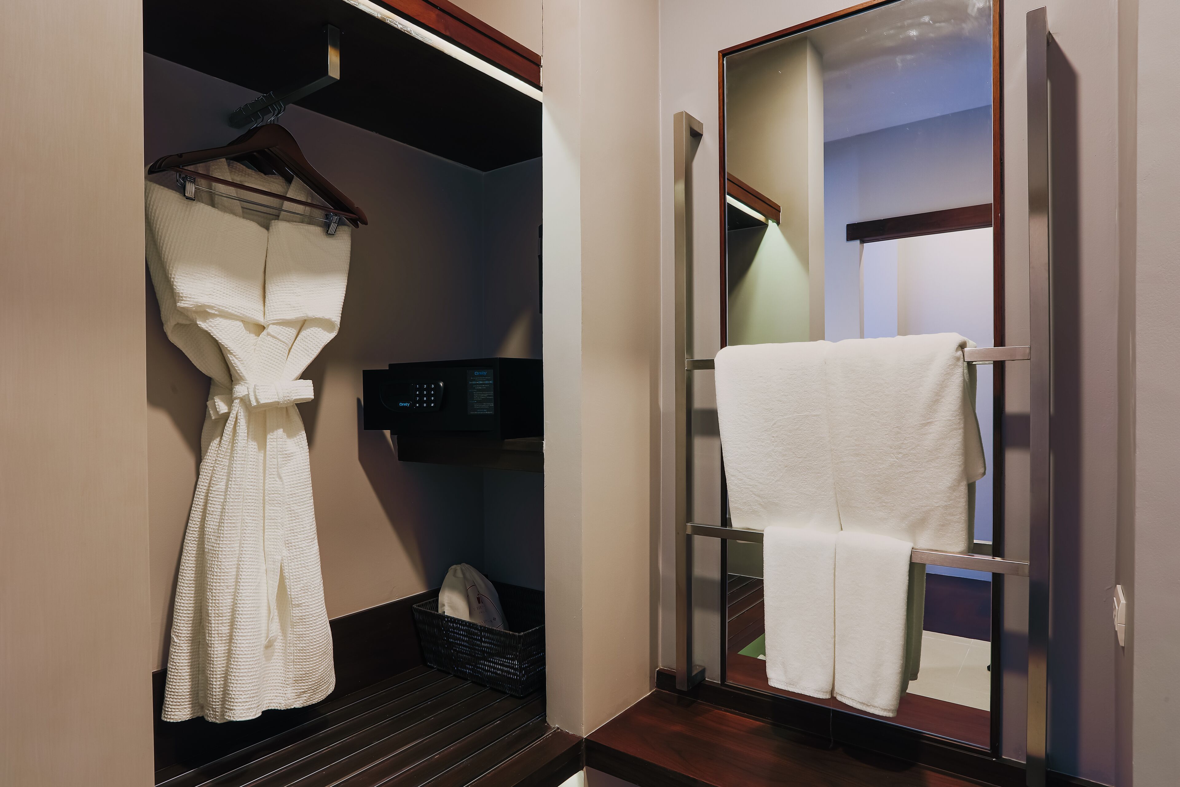 Un baño de hotel con una bata colgada en un perchero, un toallero con una toalla y una cesta negra en el suelo.