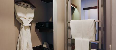 A hotel bathroom with a robe hanging on a rack, a towel rack with a towel, and a black basket on the floor.