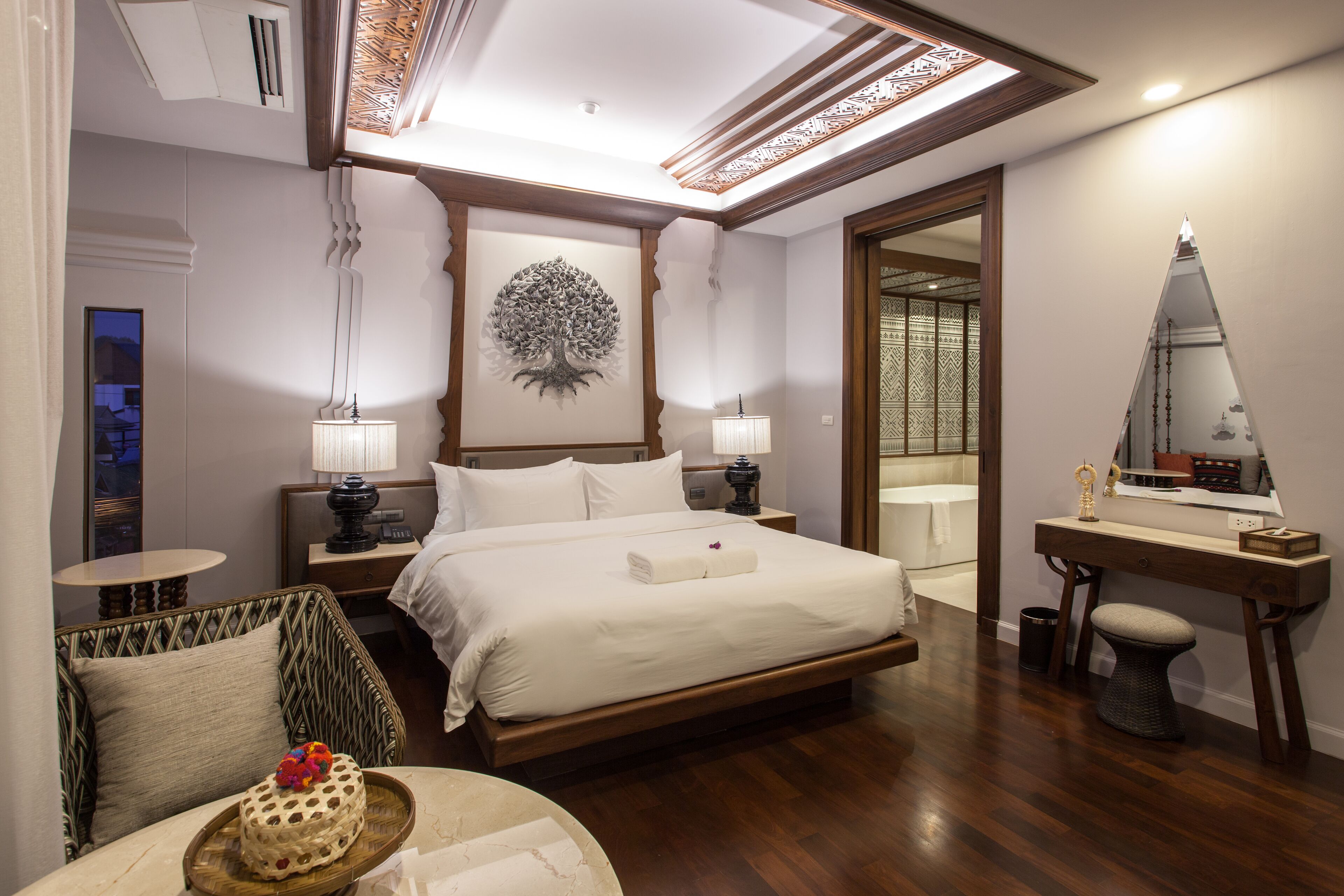 A hotel room with a large bed, bedside tables, a desk with a mirror, a small round table, and a decorative tree artwork on the wall.