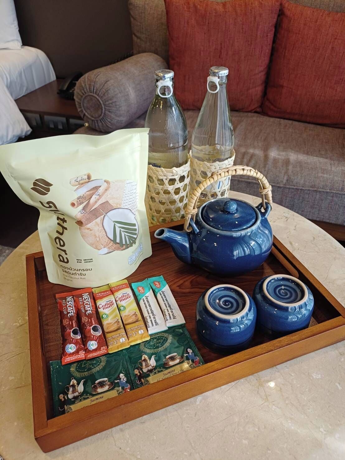 A tray with a blue teapot, two glass bottles, a bag of coffee, and several tea packets on a wooden table.