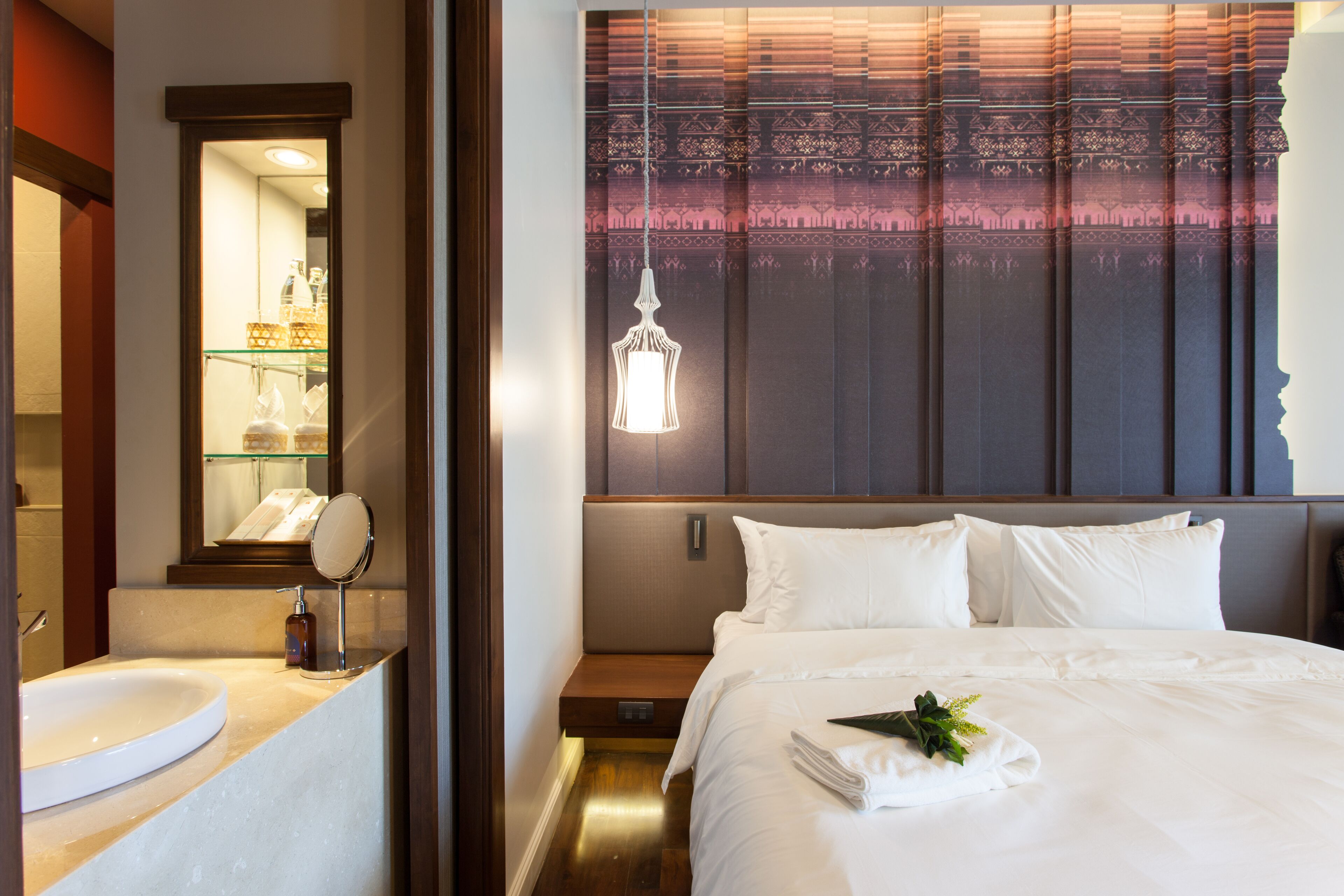 Habitación de hotel con una cama grande, una mesita de noche, un espejo y un baño con lavamanos y estantes.