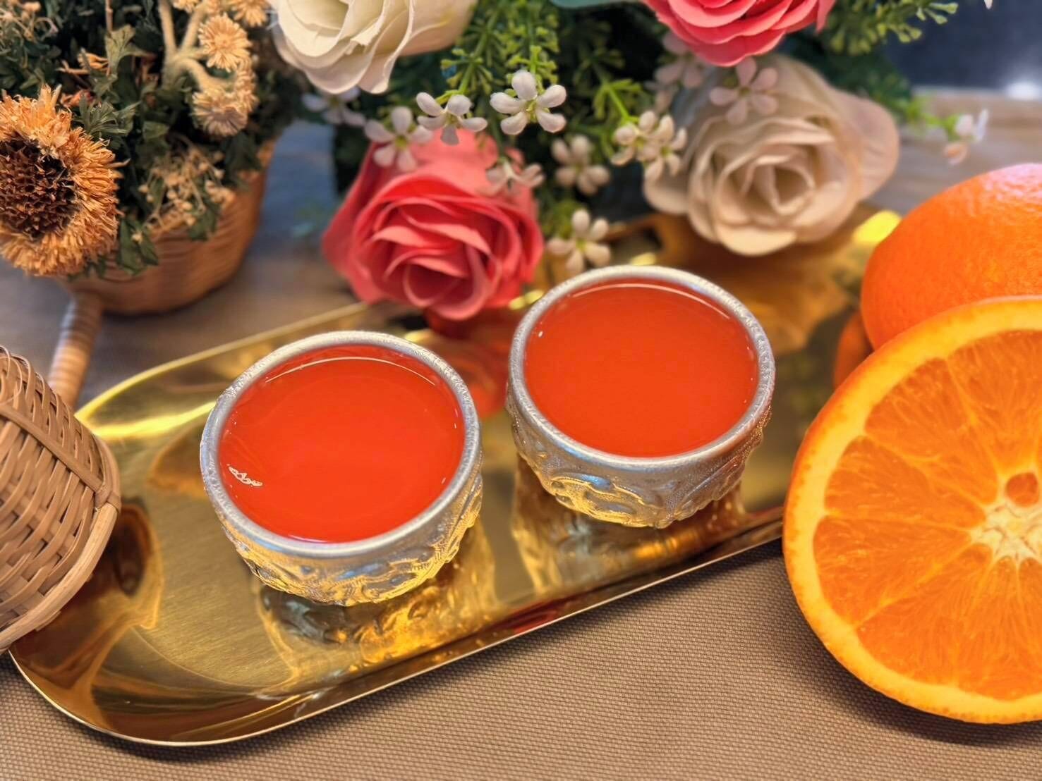 Deux verres contenant un liquide rouge sont posés sur un plateau, accompagnés d’une demi-orange et d’un bouquet de fleurs.