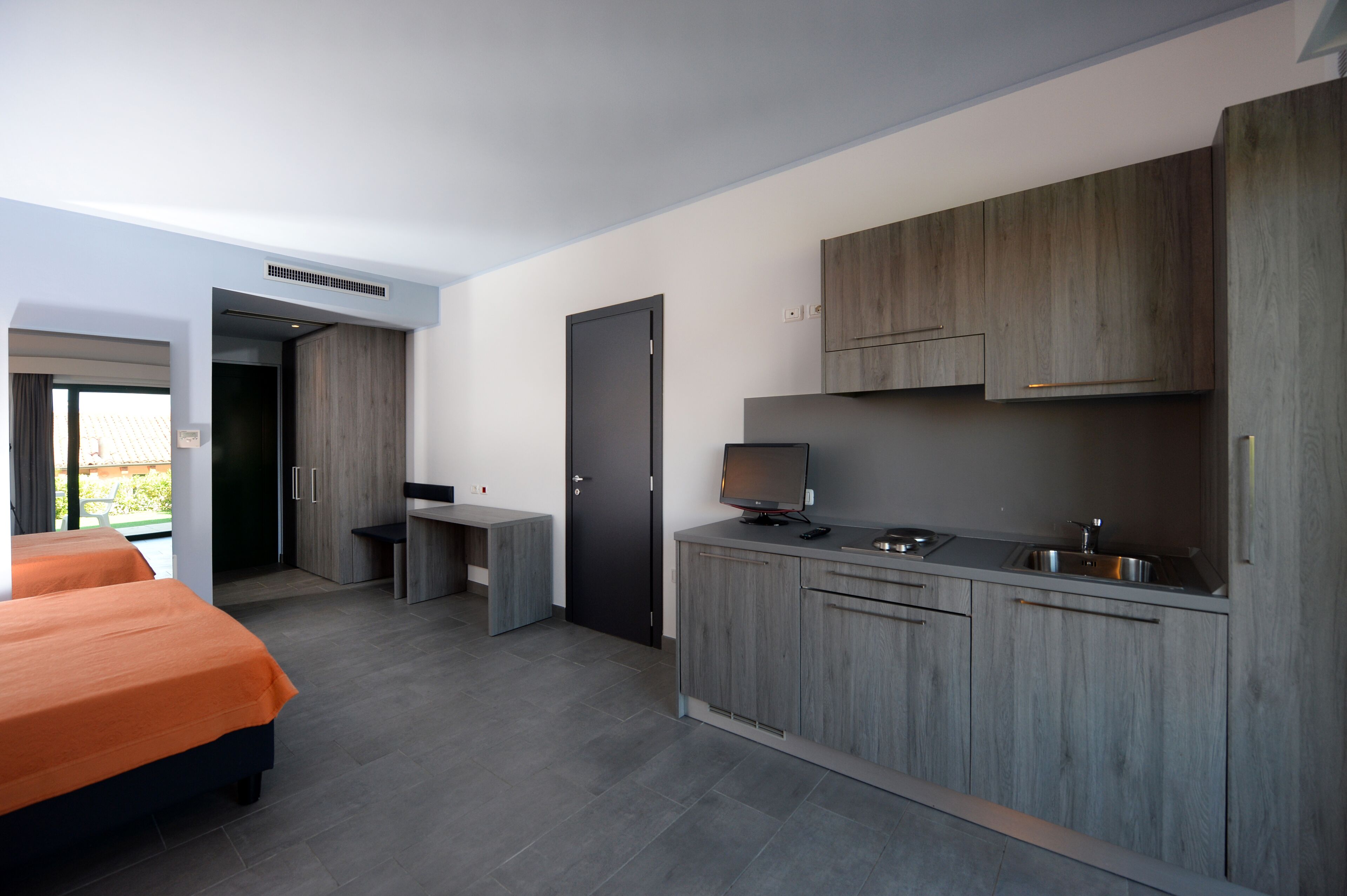 A modern hotel room with a kitchenette, a bed with orange bedding, and a view of the outdoors.