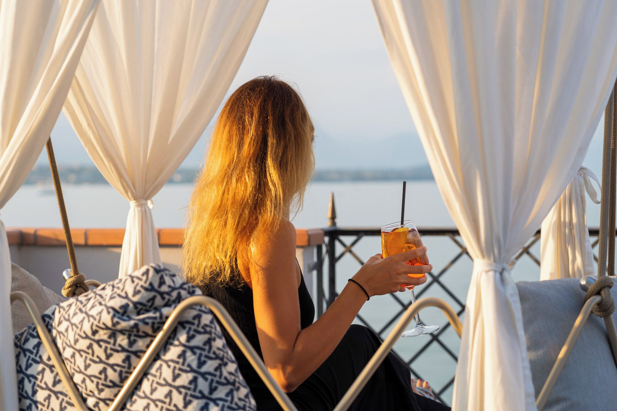 A person sitting on a balcony with a drink, overlooking a body of water.
