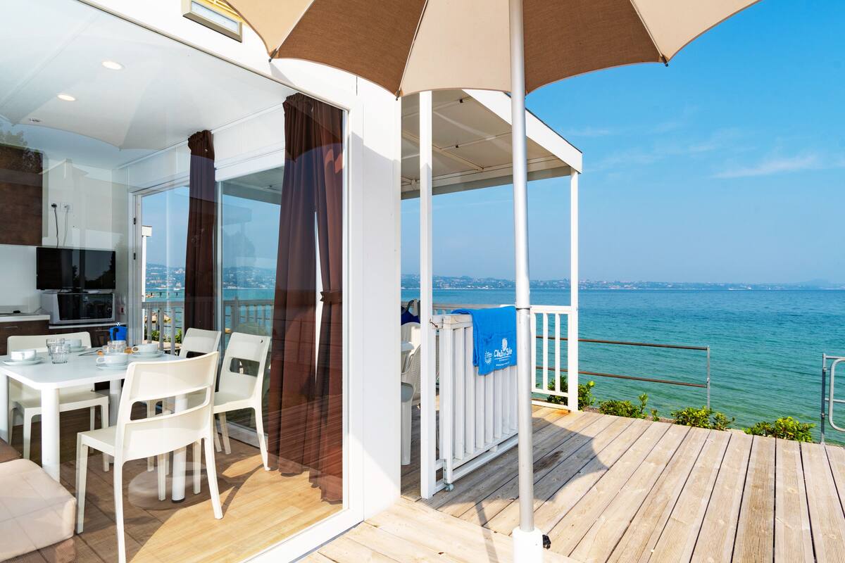 Un área de comedor moderna al aire libre con vista al mar, un espacio de estar cubierto y una cocina con televisor.