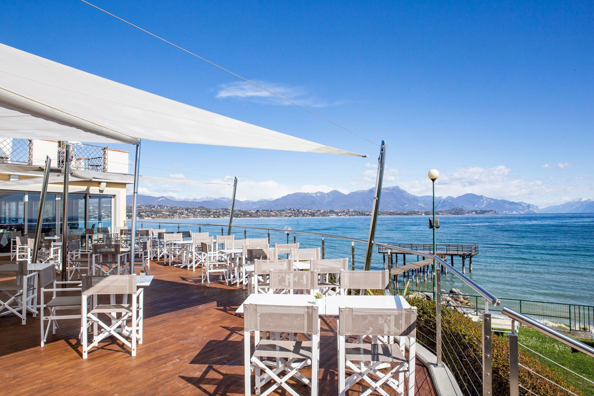 An outdoor seating area with white chairs and tables, overlooking a body of water with mountains in the background.