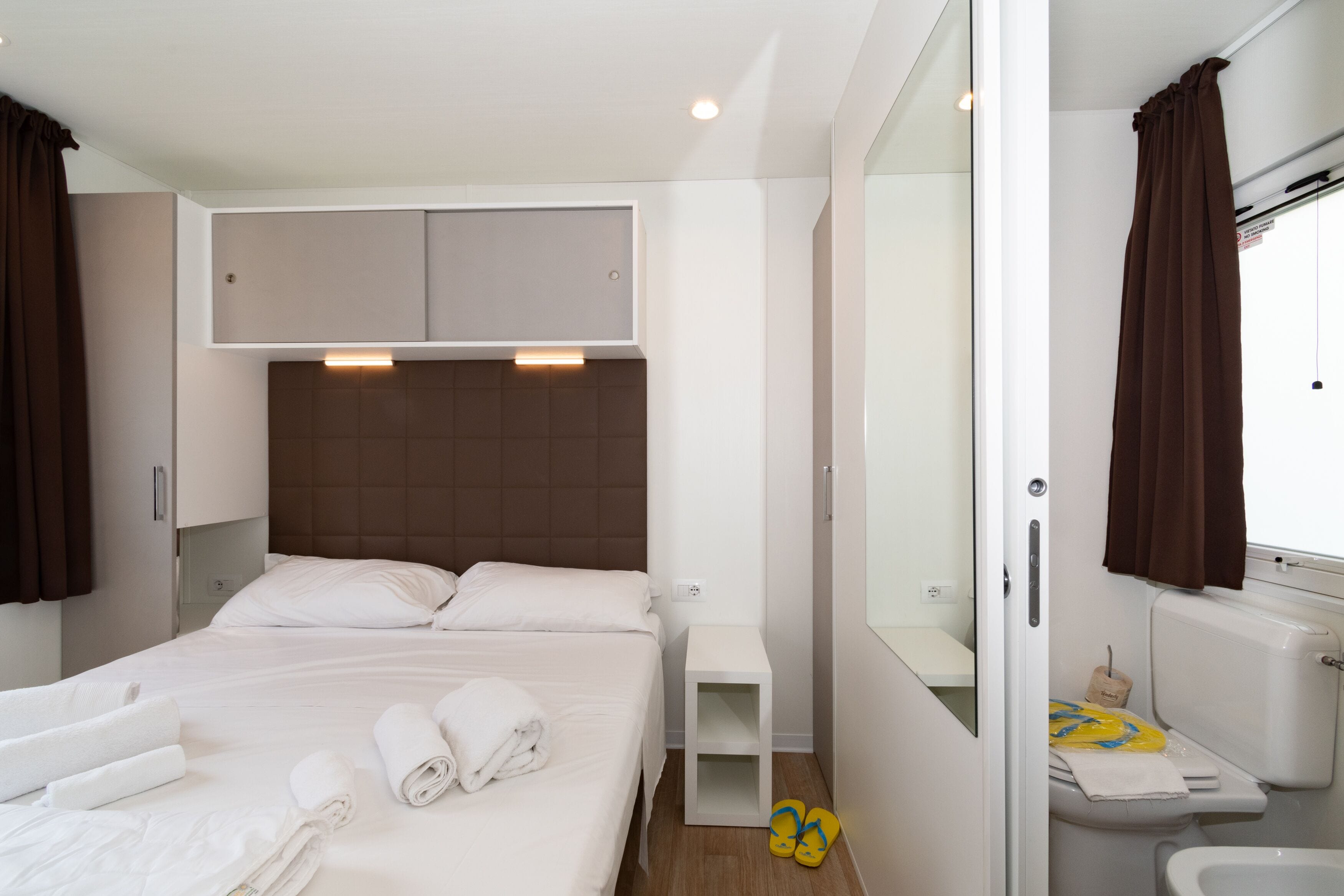 Habitación de hotel moderna con una cama grande, una mesita de noche, una pequeña zona de estar y un baño con inodoro y ducha.