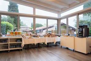 Un buffet de petit-déjeuner comprenant une machine à café, divers aliments et offrant une vue sur un quartier résidentiel grâce à de grandes fenêtres.