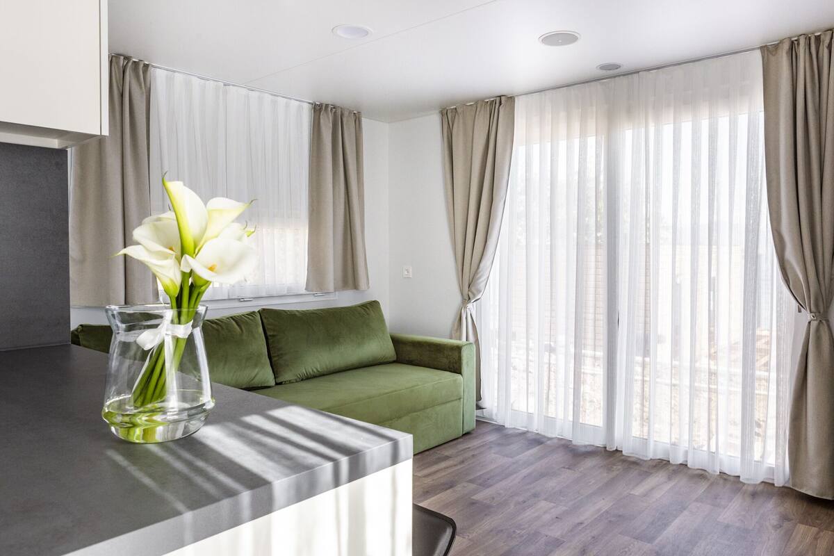 Una sala de estar moderna con un sofá verde, un jarrón transparente con flores y cortinas traslúcidas.
