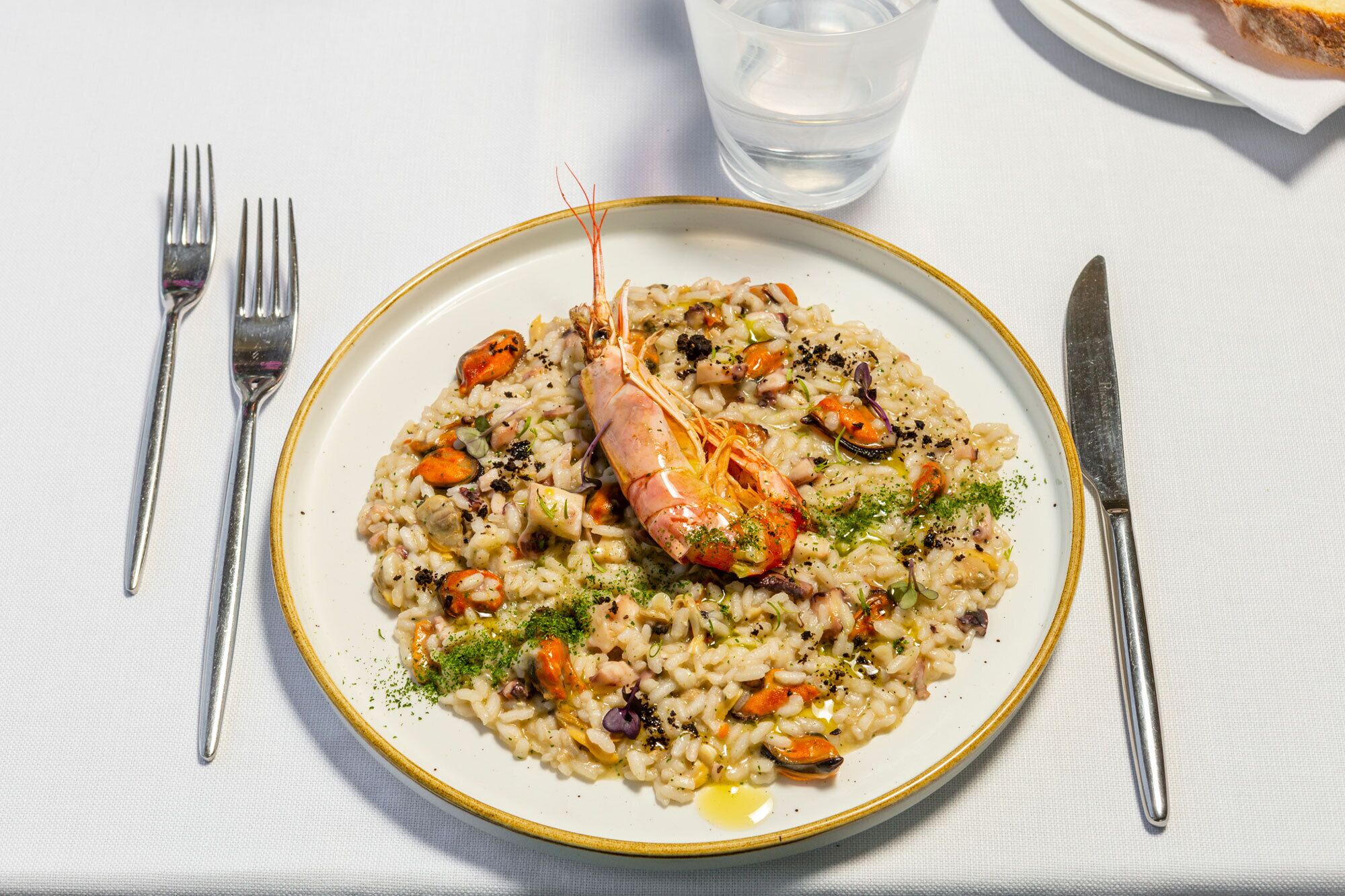 A plate of seafood risotto with a shrimp centerpiece, accompanied by cutlery and a glass of water on a white tablecloth.
