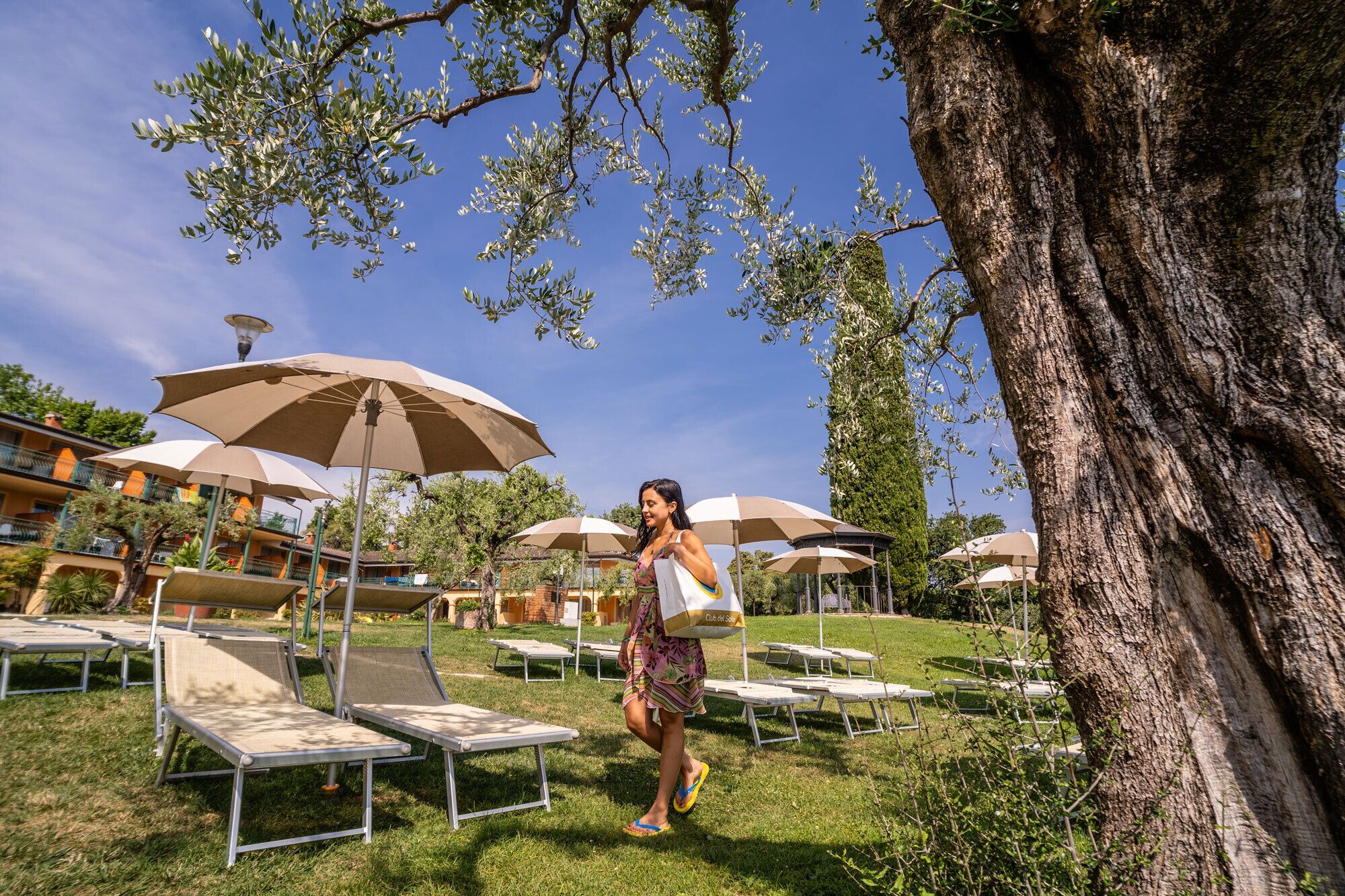 A person walking near lounge chairs and umbrellas in a sunny outdoor area with trees and buildings in the background.