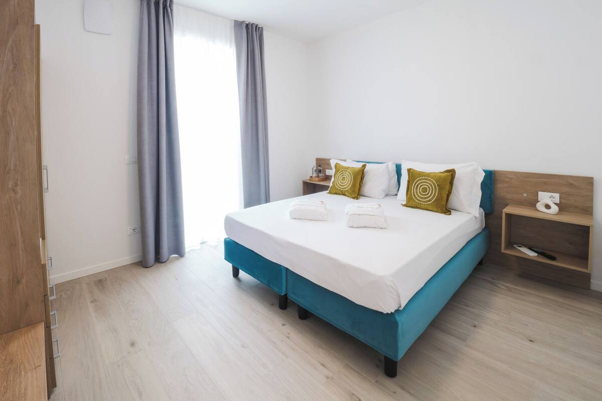 Una habitación de hotel moderna con cama, mesita de noche, cabecera de madera y ventana con cortinas.