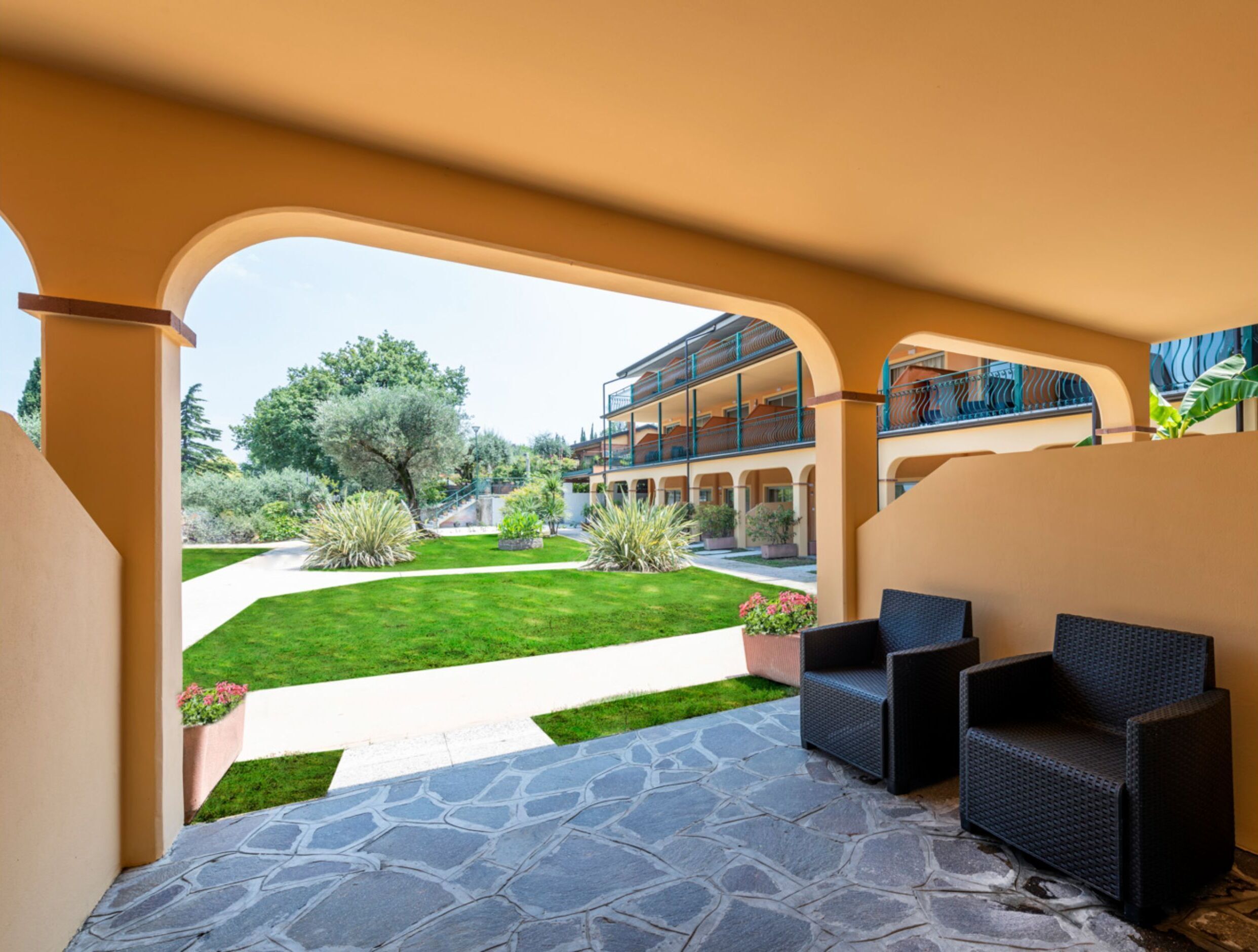 Un espacio al aire libre cubierto con suelo de piedra, dos sillas de mimbre y vistas a un edificio con balcones y un jardín.