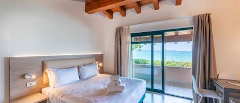 Une chambre moderne avec un grand lit, une table de chevet, un bureau et une vue sur la mer, accessible par une porte-fenêtre coulissante.