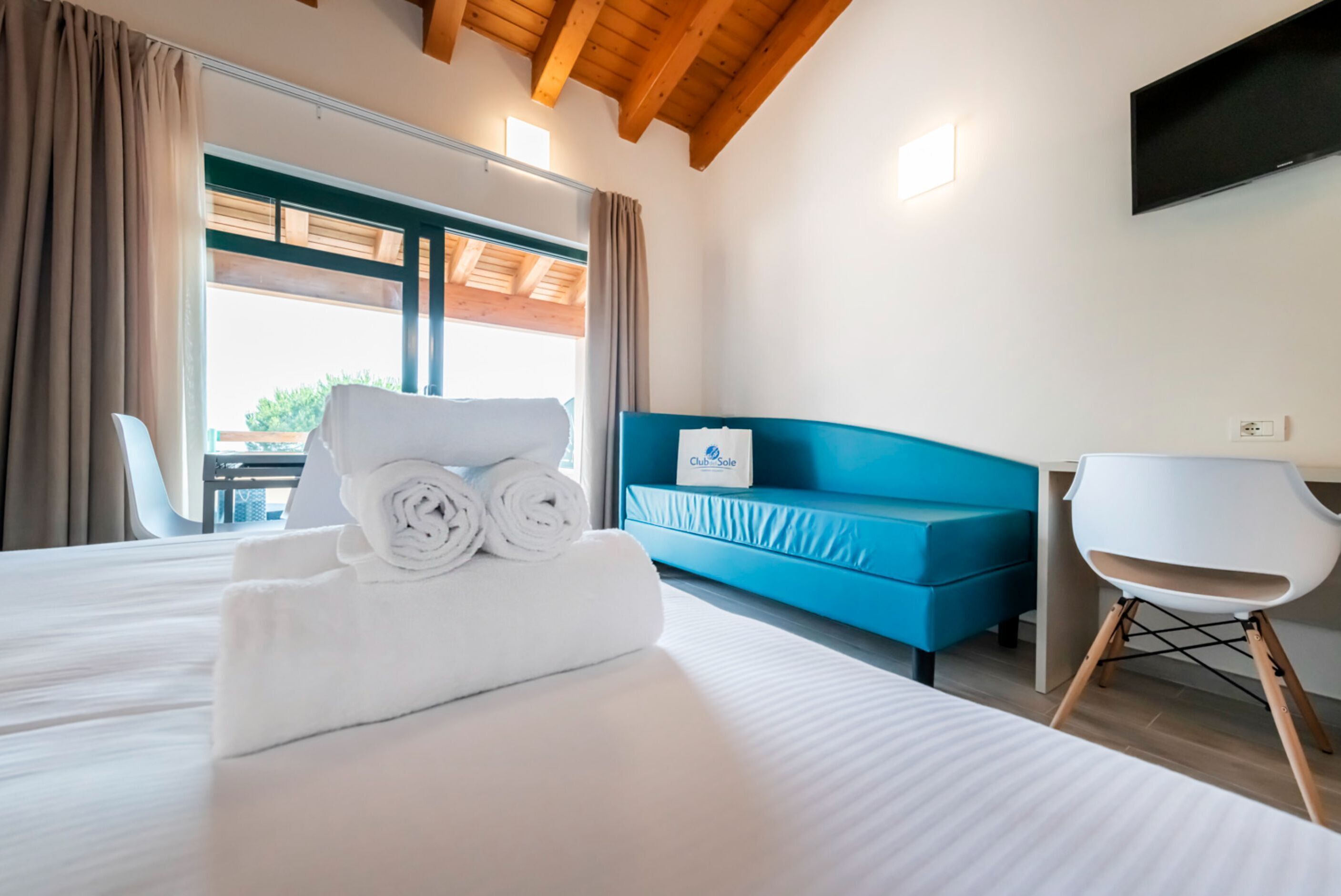 Una habitación de hotel con una cama, un sofá azul, una silla blanca y un televisor en la pared.