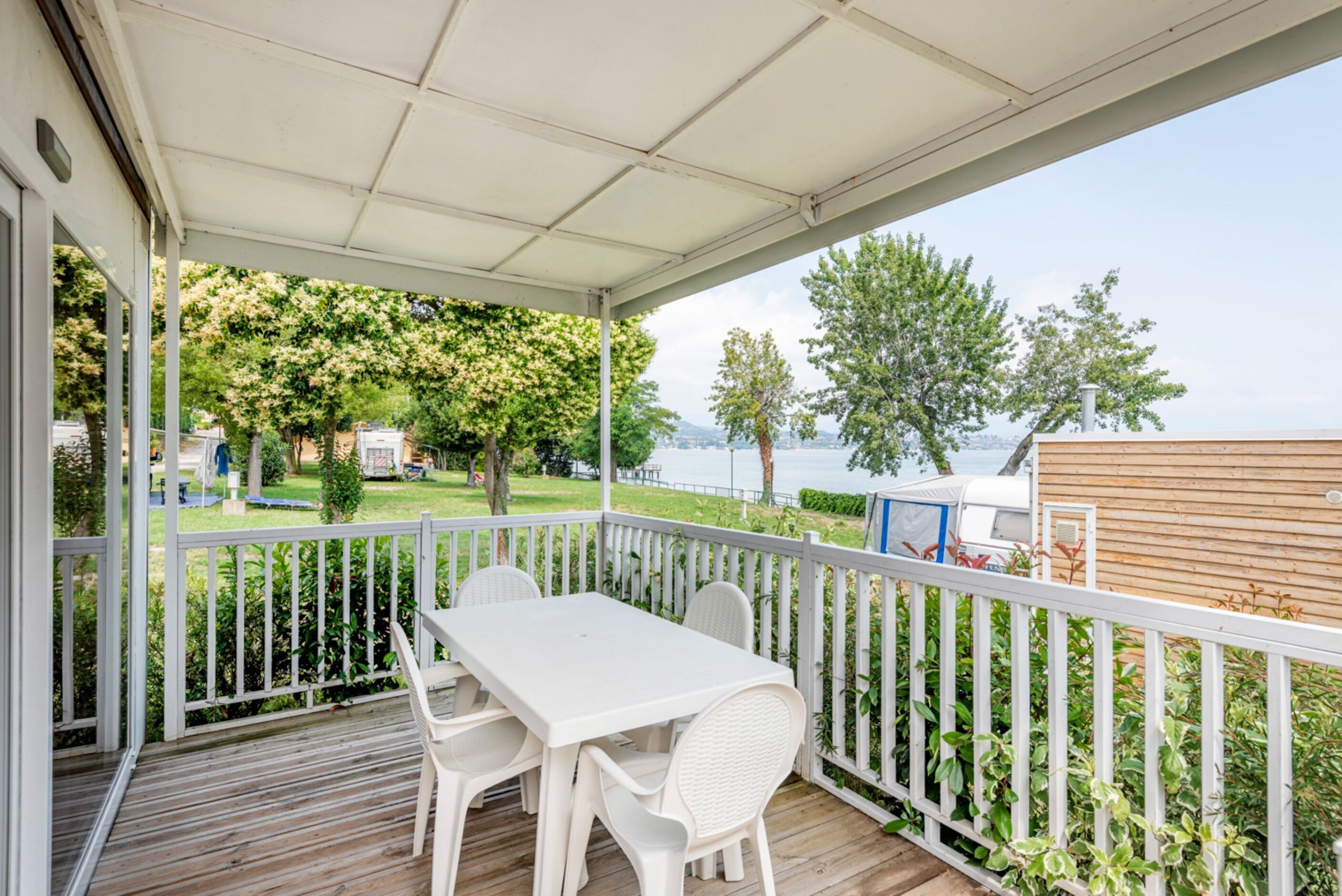 A covered outdoor area with a white table and chairs, a wooden deck, and a view of a lake and trees.
