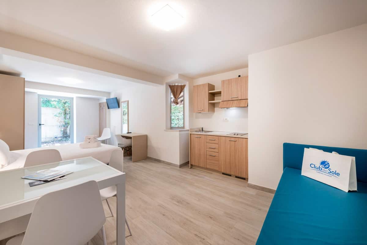 Habitación compacta con mesa de comedor, cama, kitchenette y ventana con vista a follaje.
