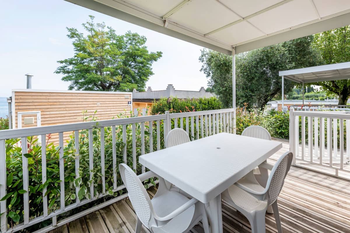 Un patio techado al aire libre con una mesa y sillas blancas, rodeado de barandas blancas, con vistas a un jardín y un edificio de fachada de madera.