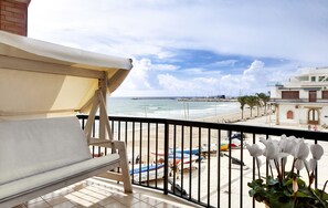 A balcony with a cushioned chair and a white canopy, overlooking a beach with boats and a coastal building. - Bright apartment overlooking the beach and direct access to the sea (Marina di Ragusa)