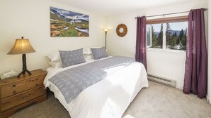 Ein Schlafzimmer mit einem Bett, einem Nachttisch, einer Lampe, einem an der Wand befestigten Kranz und einem Fenster mit Vorhängen.