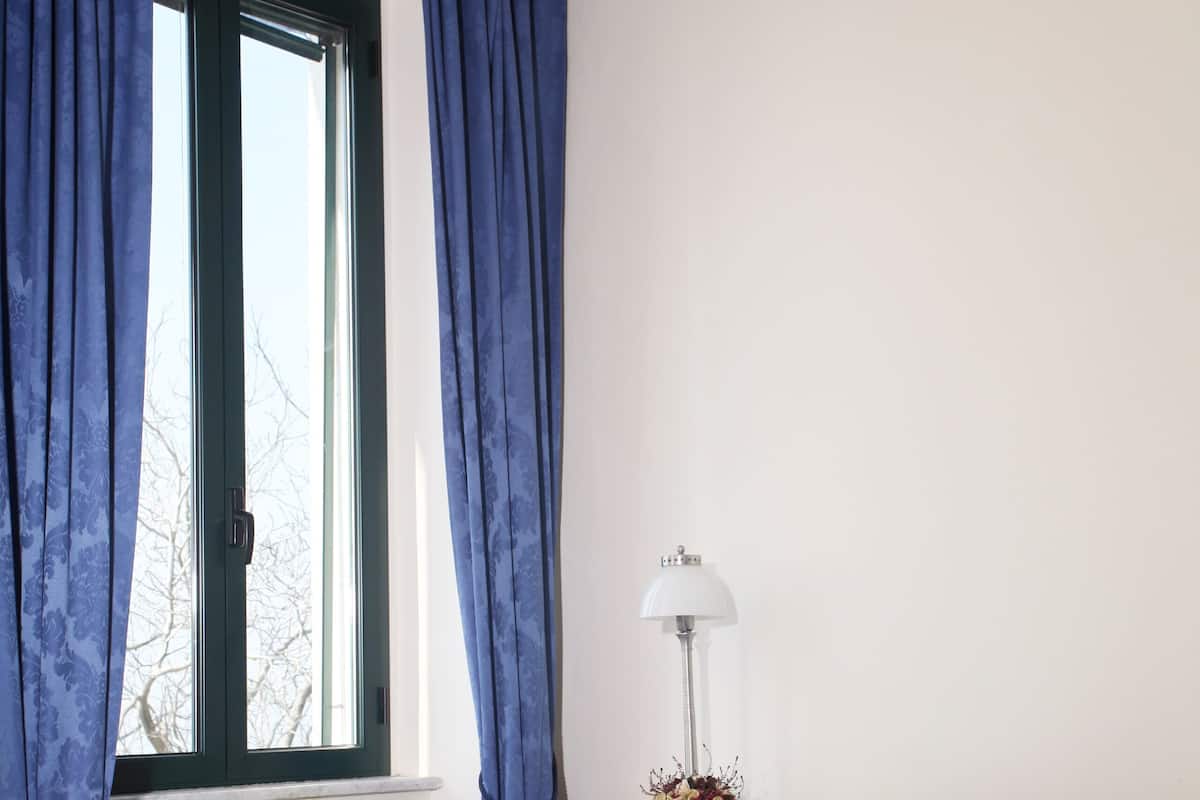 A bedroom with a large bed, blue curtains, a window, and a bedside table with a lamp.