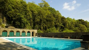 A swimming pool with a stone wall and arched doorways, surrounded by lush greenery. - Villa Palazzola (Albano Laziale)