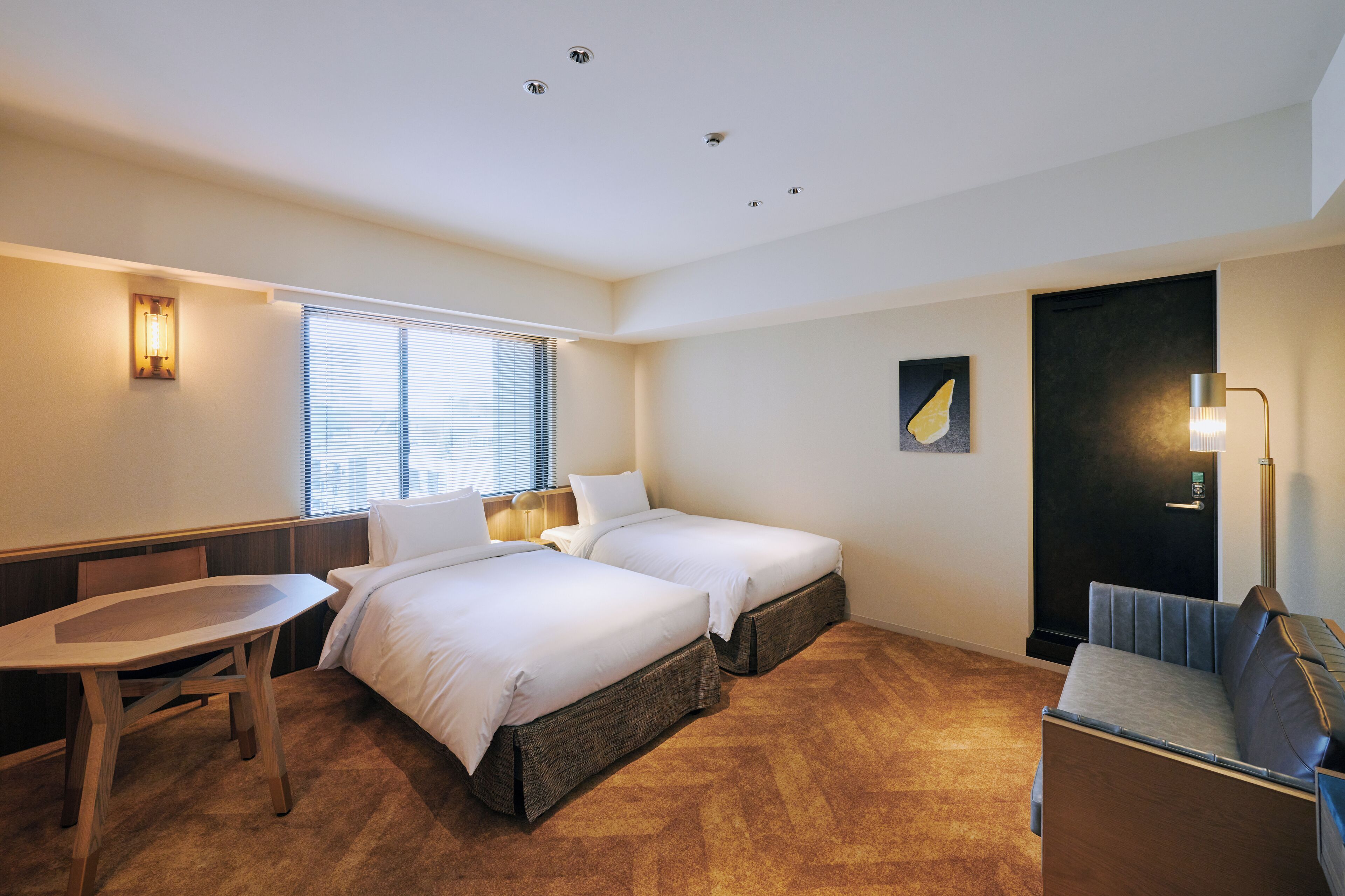 Habitación de hotel con dos camas, un escritorio, una silla, un sofá y una ventana con persianas.