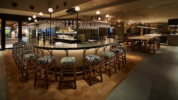 Zona de bar con barra circular, varios taburetes y copas colgantes.