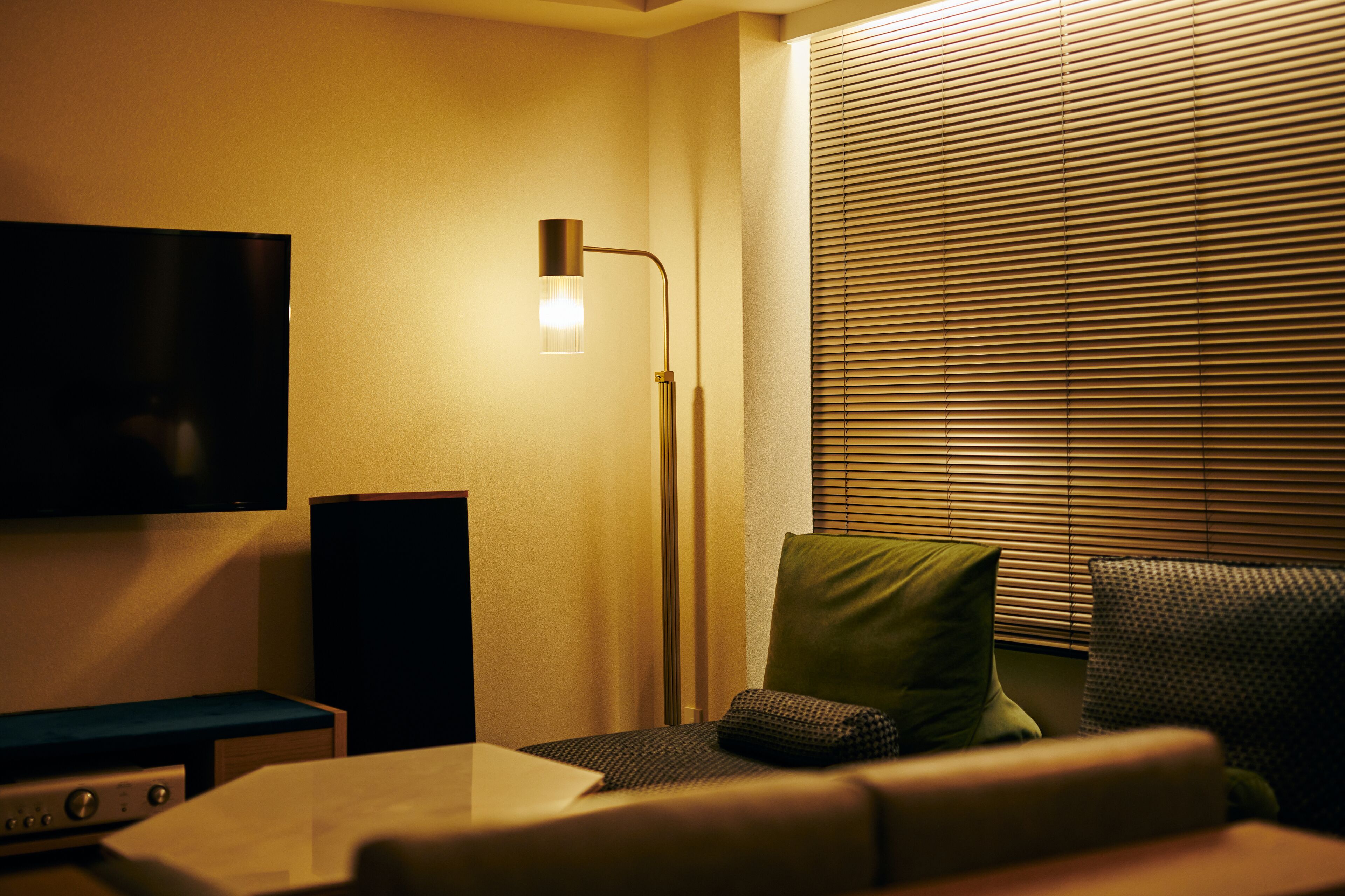 A hotel room with a flat-screen TV mounted on the wall, a standing floor lamp, a sofa with cushions, and a window with blinds.