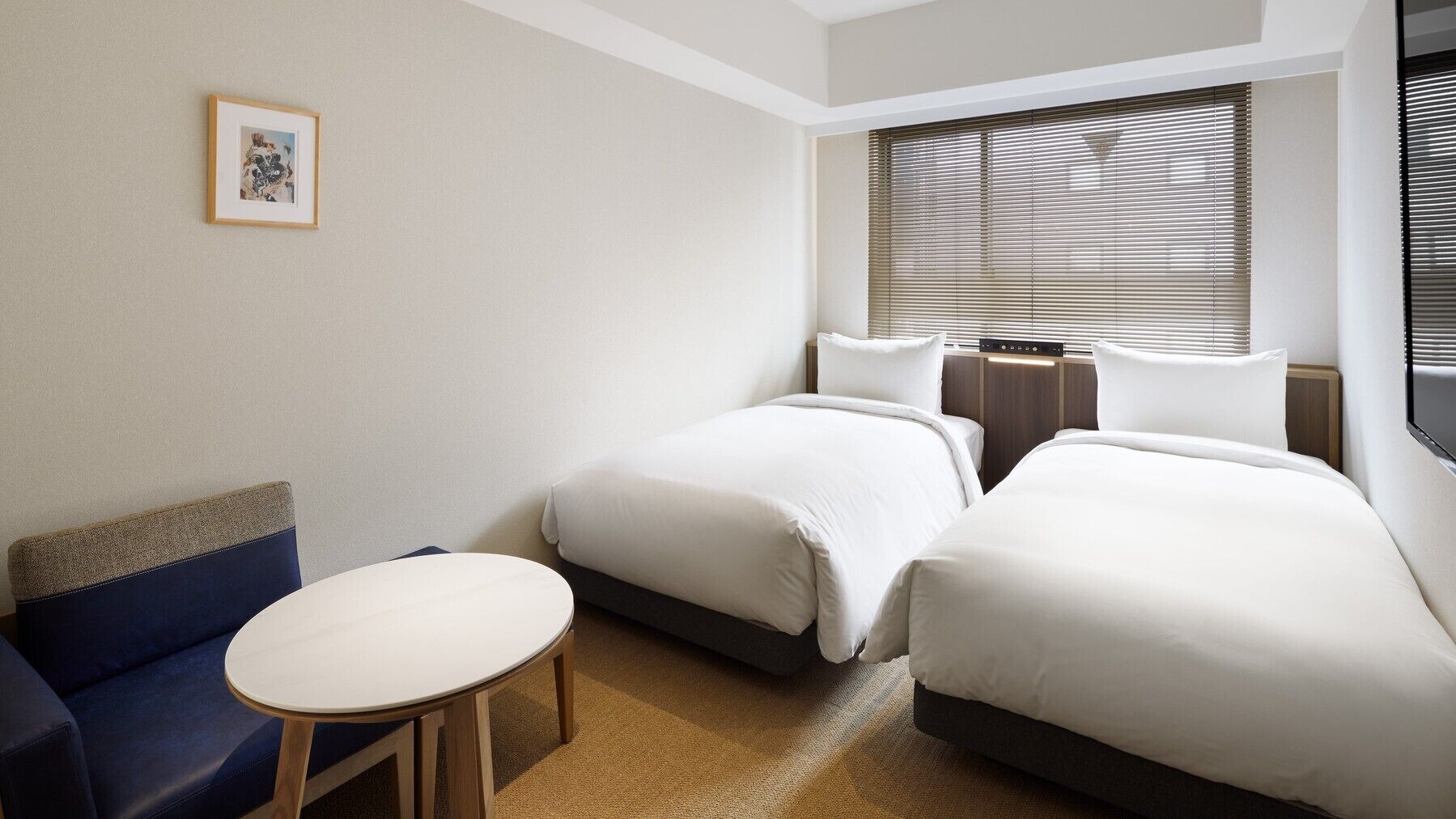 A hotel room with two beds, a small table, a blue sofa, and a window with blinds.
