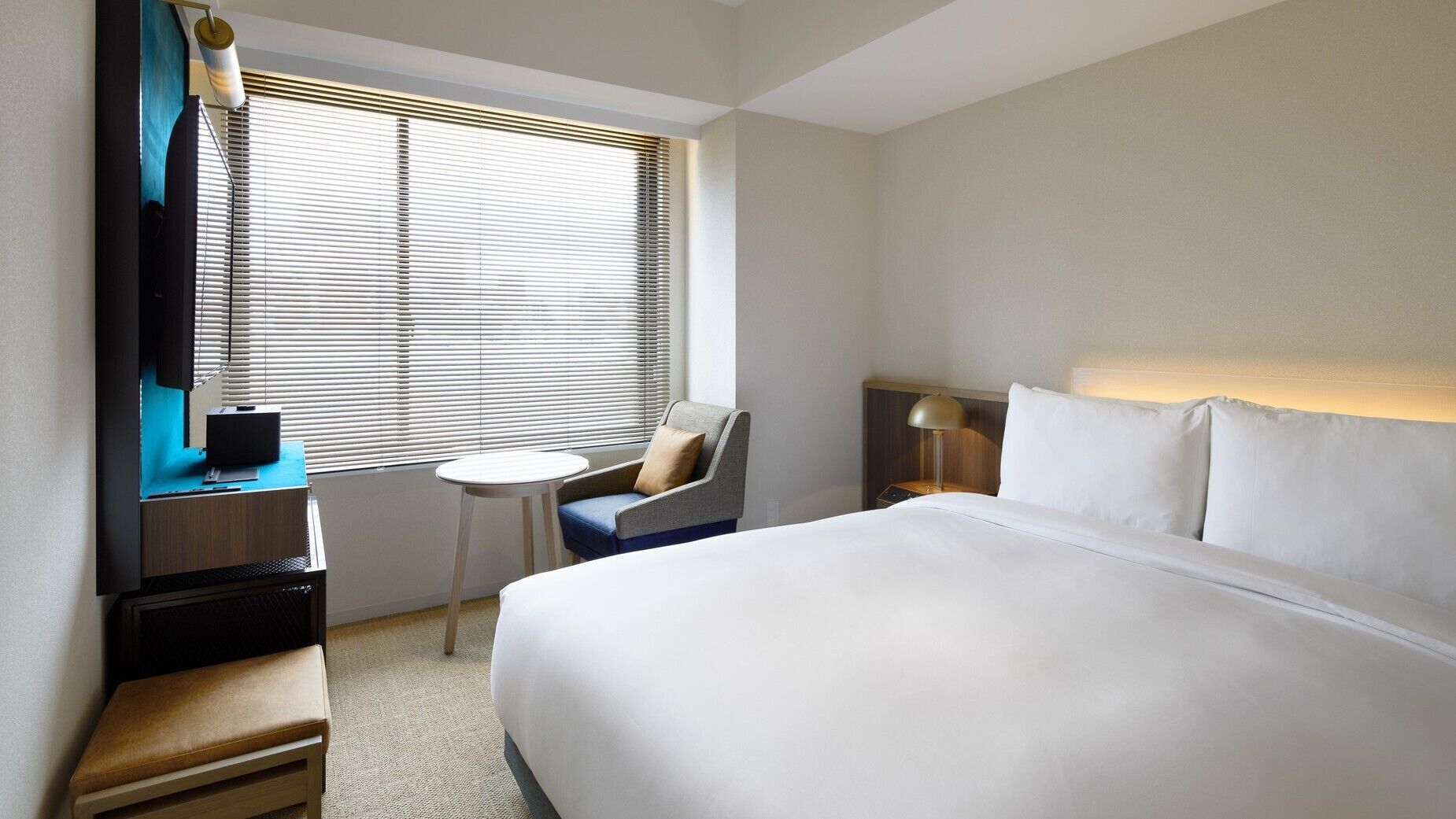 A hotel room with a bed, a desk, a chair, a small table, and a window with blinds.