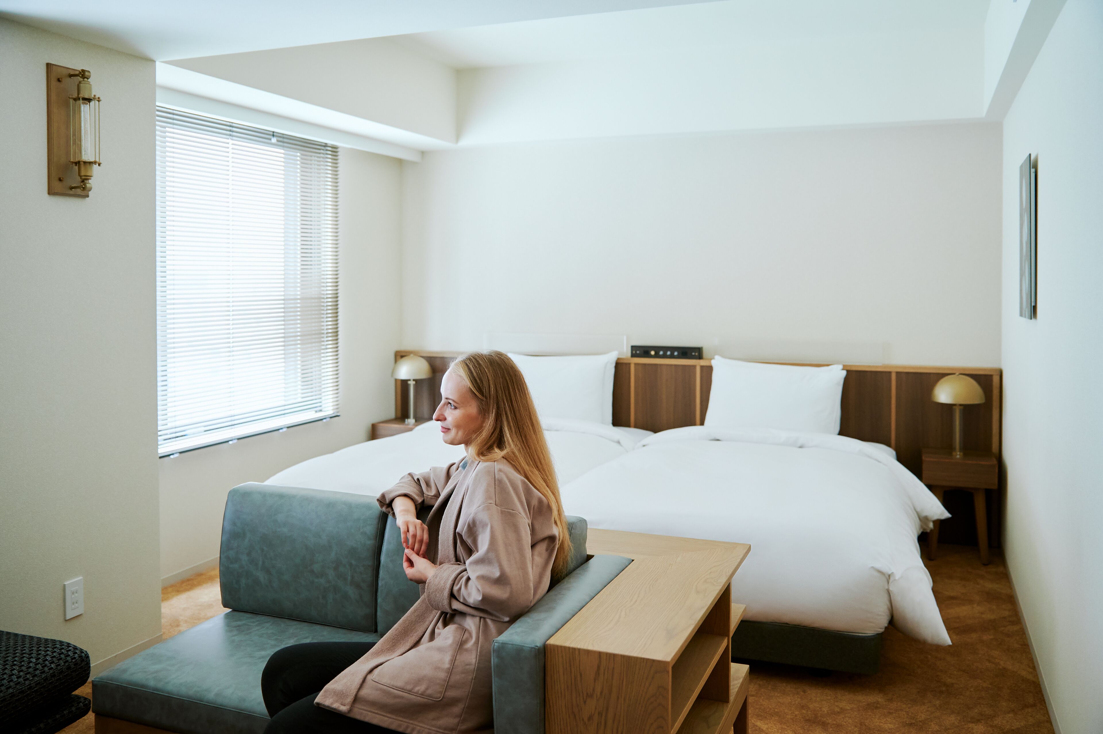 A hotel room with a bed, a nightstand, a lamp, a window with blinds, and a seating area with a bench.