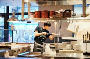 A kitchen with stainless steel appliances, a person working at a counter, and various kitchen utensils and dishes.