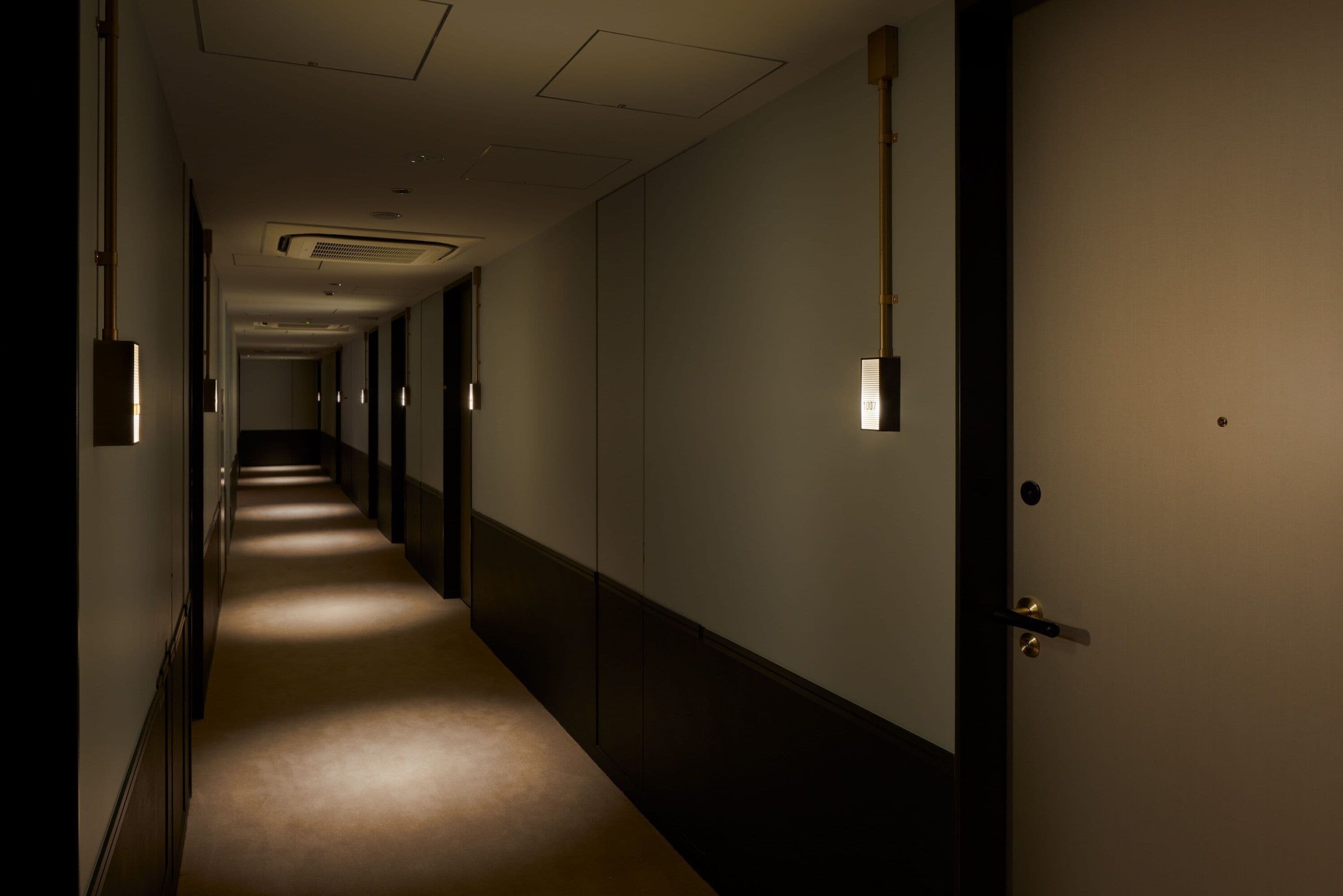 Een lange, smalle hotelgang met vloerbedekking, een deur aan de rechterkant en wandlampen.