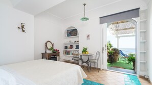 A bedroom with a bed, a desk, a chair, a small table, and a view of the sea through an open door. - Elia's room: Double room with bathroom and shared terrace with sea view (Praiano)