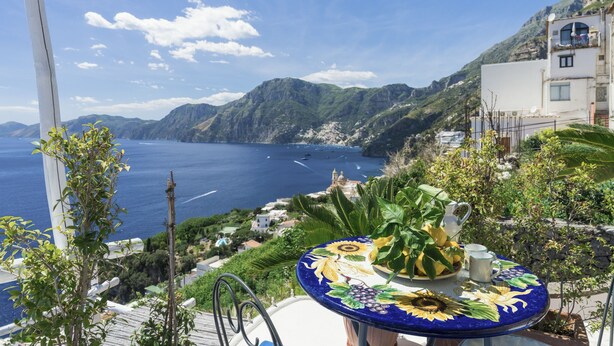 A table with a floral design, a pitcher, and a cup on a balcony overlooking a coastal landscape with mountains and a sea view. - Elia's room: Double room with bathroom and shared terrace with sea view (Praiano)