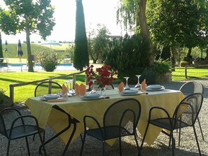 An outdoor dining area with a table set for four, surrounded by chairs, in a garden setting with a pool and trees in the background. - Rosa dei Venti (Cortona)